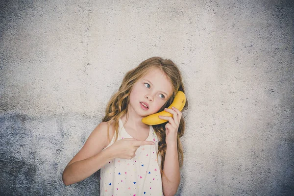 灰色のコンクリートの壁の背景にバナナとの電話をしているきれいな女の子 — ストック写真