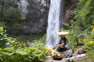European man with beard and japanese robe making fire ceremony in front of Great Waterfall in Austria clipart