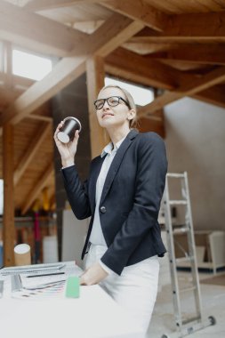 Siyah ceketli genç bayan mimar elinde kahve fincanı ve evin çatısındaki planlarla masanın yanında poz veriyor.