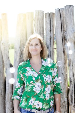 yaz güneşli bir günde ahşap çit yakınında uçan sabun kabarcıkları arasında poz mutlu sarışın kadın 