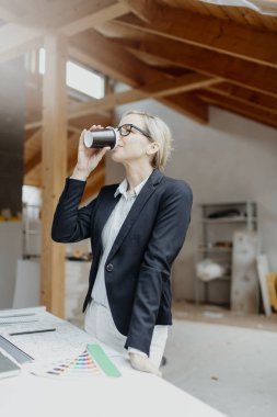Siyah ceketli genç bayan mimar elinde kahve fincanı ve evin çatısındaki planlarla masanın yanında poz veriyor.