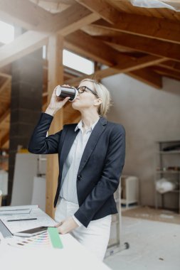 Siyah ceketli genç bayan mimar elinde kahve fincanı ve evin çatısındaki planlarla masanın yanında poz veriyor.