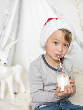 şişe tutan ve saman evde süt içmek kırmızı santa claus şapka giyen çocuk 
