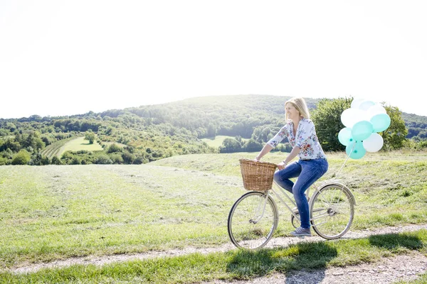 Happy woman riding bicycle with balloons on summer meadow at sunny day