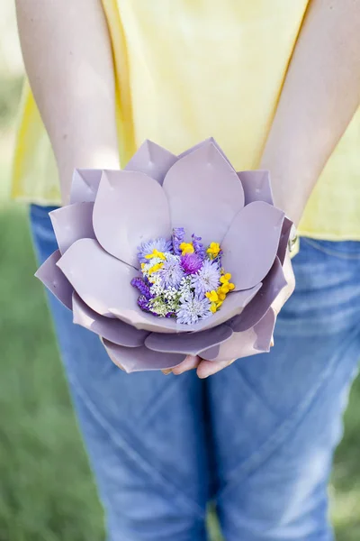 牧草地で花と美しい紫色の花のボウルを保持する女性の手 — ストック写真