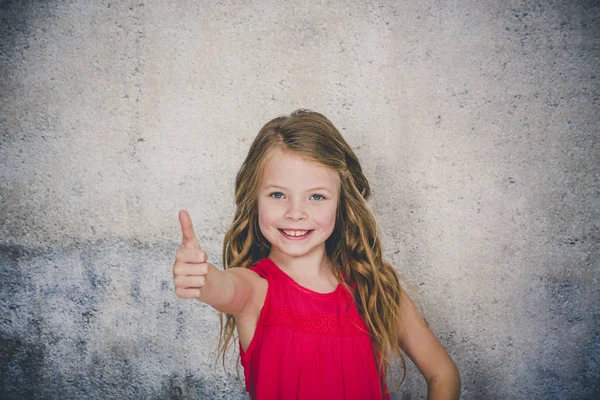 cute blonde girl in red blouse showing thumb up and looking at camera in front of concrete background