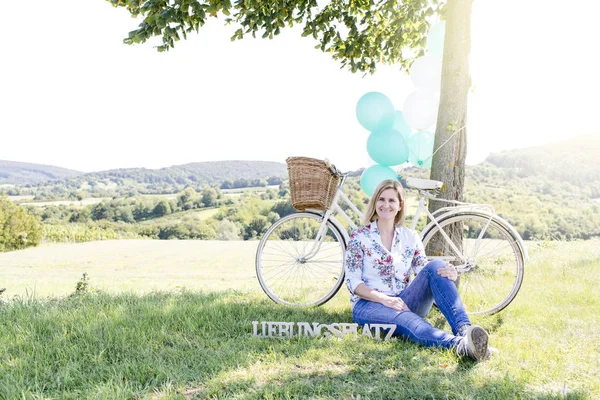 Happy woman sitting on green grass with wooden lettering chase in German near bicycle and balloons with tree on summer meadow