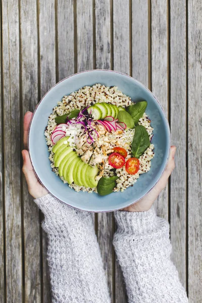 hands holding Healthy Quinoa Bulgur bowl with vegetables and chicken over wooden background