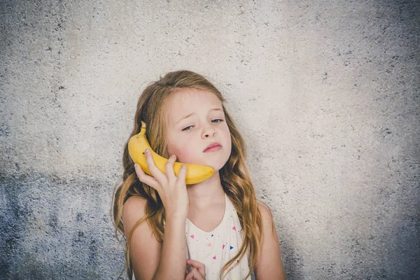 灰色のコンクリートの壁の背景にバナナとの電話をしているきれいな女の子 — ストック写真