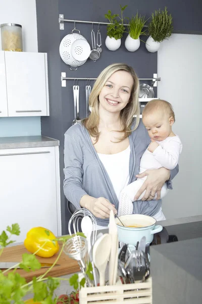 happy mother holding little baby and cooking in kitchen at home