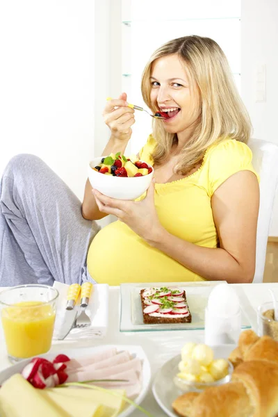 椅子に座っていると家庭での料理とテーブルの近くに座ってボウルからフルーツ サラダを食べて幸せな妊娠中の女性 — ストック写真