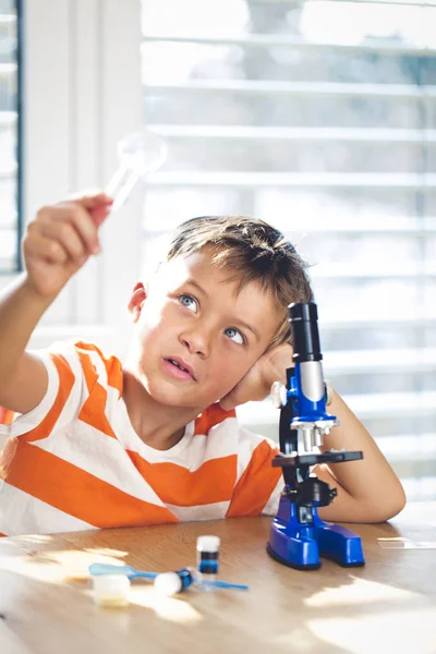 顕微鏡と科学実験をしながら虫眼鏡を通して見る近くのテーブルに座っている少年 — ストック写真