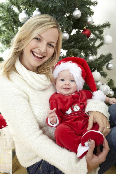 Eğleniyor kucaklayan şirin bebek iken kırmızı Noel Baba kostüm dekoratif Noel ağacı yakınındaki anne  