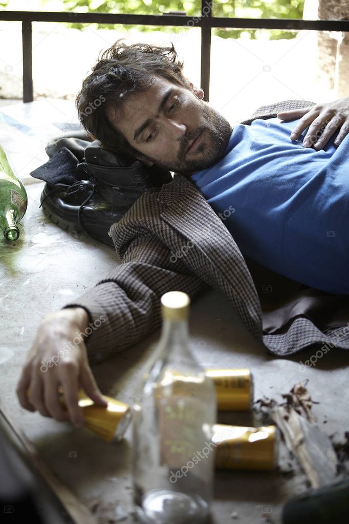 drunkard in dirty clothes lying on floor near empty alcohol bottles in neglected building 