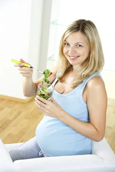 肘掛け椅子に座って 家でガラスからサラダを食べる幸せな妊娠中の女性 — ストック写真
