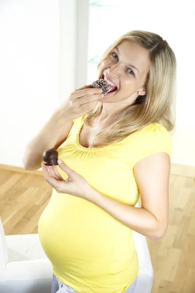 吃瑞典巧克力炸弹的愉快的怀孕的妇女站在扶手椅在家附近 — 图库照片