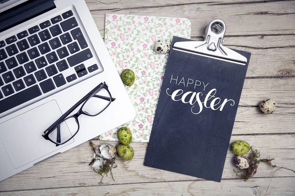 laptop with easter eggs and clipping board with text Happy Easter over light wooden background