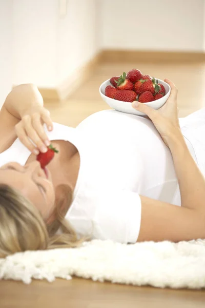 躺在地毯和吃草莓从碗在家里的愉快的怀孕妇女 — 图库照片