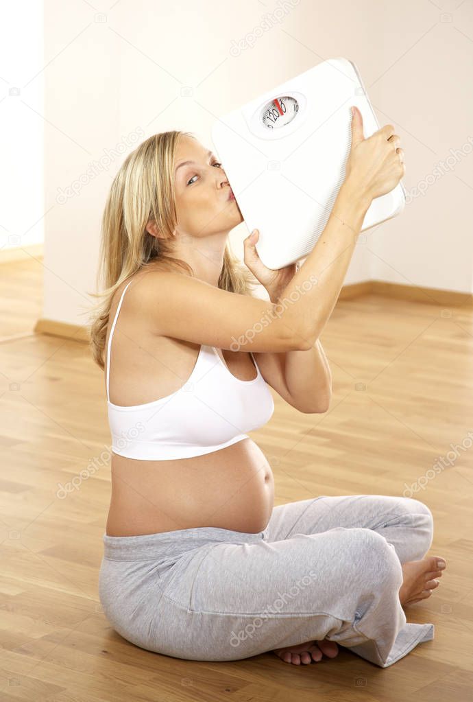 blonde pregnant woman kissing scales while sitting on floor at home 