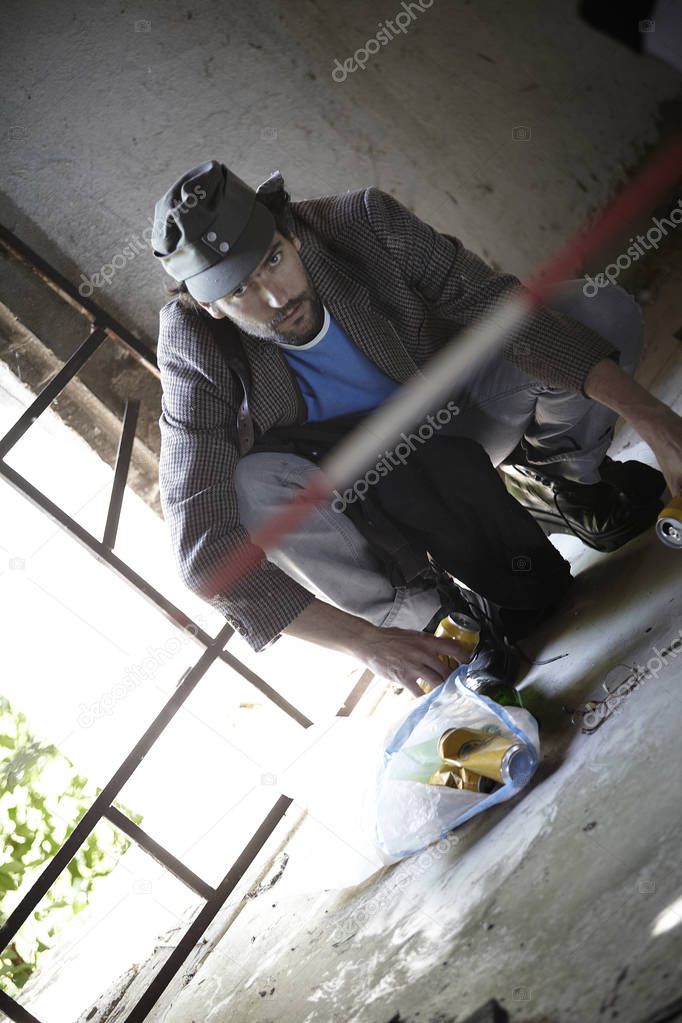 bearded homeless man squatting and picking up empty bottles from floor in old building 