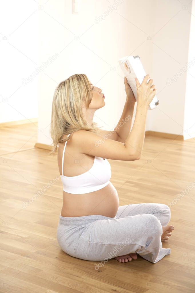 blonde pregnant woman kissing scales while sitting on floor at home 