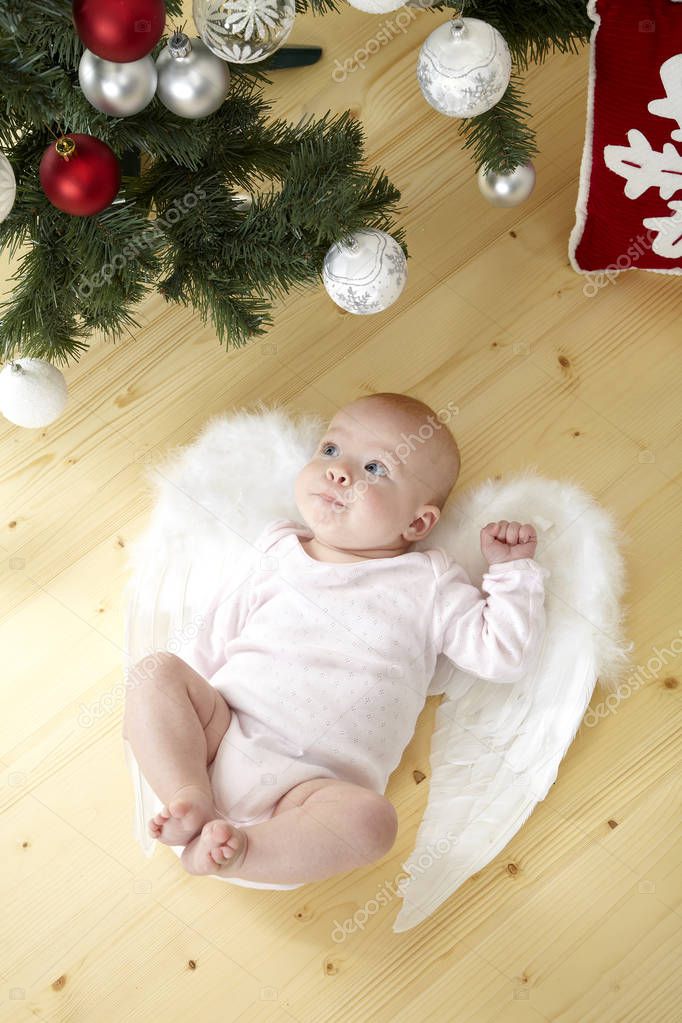 funny little baby in white costume with angel wings lying on floor near Christmas tree with baubles 
