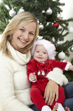 Eğleniyor kucaklayan şirin bebek iken kırmızı Noel Baba kostüm dekoratif Noel ağacı yakınındaki anne  