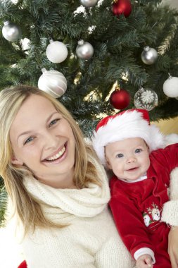 Eğleniyor kucaklayan şirin bebek iken kırmızı Noel Baba kostüm dekoratif Noel ağacı yakınındaki anne  