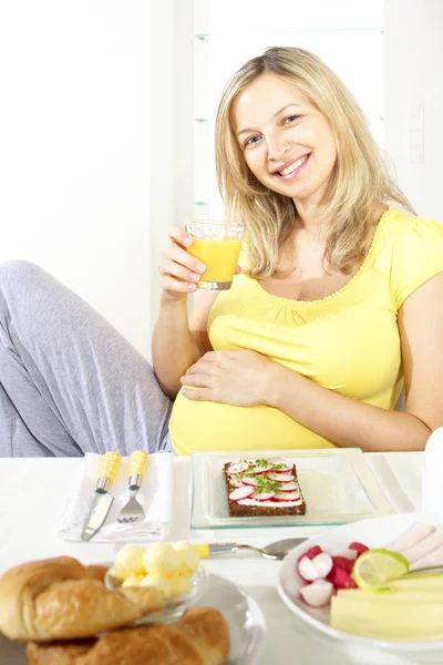 オレンジ ジュースを飲みながら自宅の料理とテーブルの近くの椅子に座って幸せな妊娠中の女性 — ストック写真