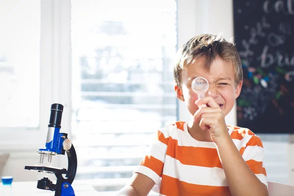 顕微鏡と科学実験をしながら虫眼鏡を通して見る近くのテーブルに座っている少年 — ストック写真