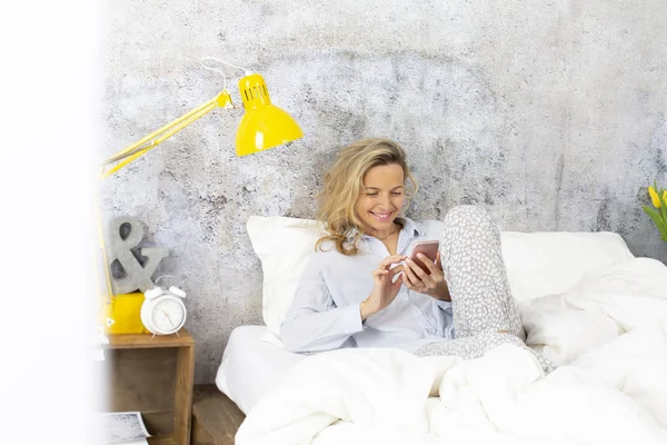 cute woman wearing pajamas reading news in smartphone while lying in bed at bedroom