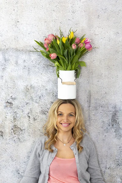 happy young woman with vase full of colorful tulips on head leaning on concrete wall and smiling , Mother day concept