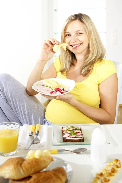 家庭での料理とテーブルの近くの椅子に座りながらチーズを食べて幸せな妊娠中の女性 — ストック写真