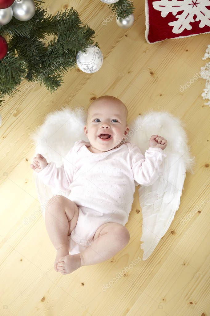 funny little baby in white costume with angel wings lying on floor near Christmas tree with baubles 