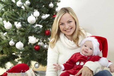 Eğleniyor kucaklayan şirin bebek iken kırmızı Noel Baba kostüm dekoratif Noel ağacı yakınındaki anne  