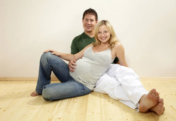 自宅で木製の床に座っている間に妊娠中の妻の腹を抱きしめている夫 — ストック写真