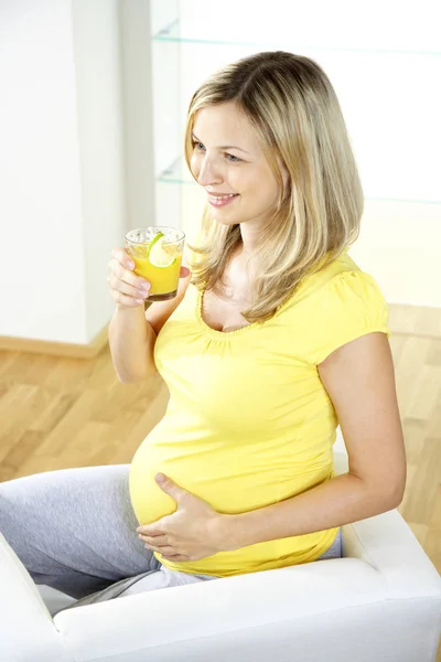 肘掛け椅子に座っていると 自宅のガラスからオレンジ ジュースを飲んで幸せな妊娠中の女性 — ストック写真