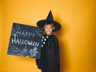 küçük boy gülümseyerek sihirbaz şapka ve mutlu halloween kara tahta tutarak ve kameraya turuncu arka plan üzerinde seyir siyah pelerin giymiş