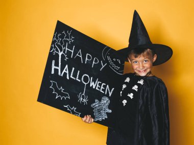 küçük boy gülümseyerek sihirbaz şapka ve mutlu halloween kara tahta tutarak ve kameraya turuncu arka plan üzerinde seyir siyah pelerin giymiş