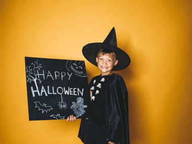 küçük boy gülümseyerek sihirbaz şapka ve mutlu halloween kara tahta tutarak ve kameraya turuncu arka plan üzerinde seyir siyah pelerin giymiş