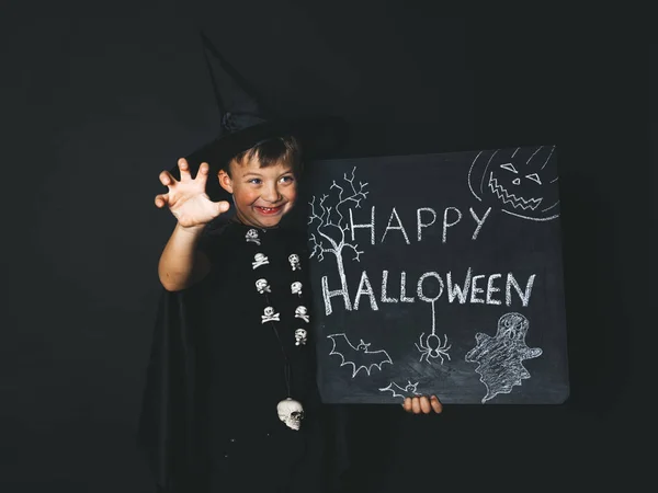 smiling boy dressed magician costume and black cape holding happy halloween chalkboard and showing frightening gesture while looking aside