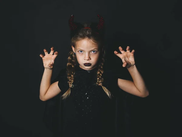 little girl in halloween devil costume with horns on head looking at camera with frightening gesture on black background