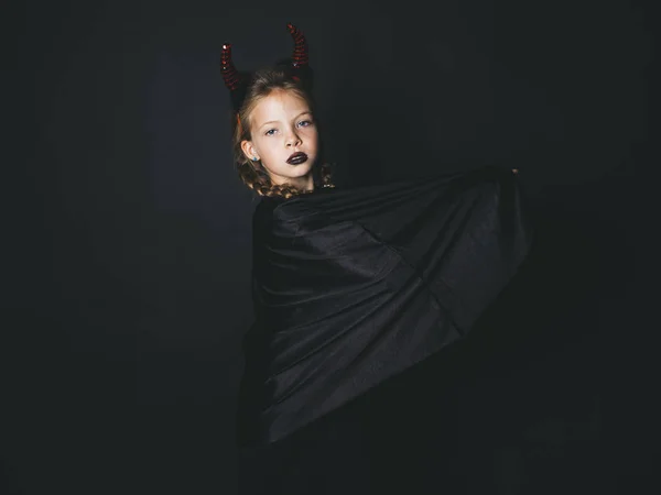 小女孩在万圣节魔鬼服装与头上的角和黑色斗篷看着相机 而在黑色背景上摆出 — 图库照片