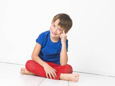 dalgın altı yaşında bir çocuk kırmızı pantolon ve gülümseyen ve çapraz ayaklı beyaz katta otururken kameraya bakarak mavi t-shirt