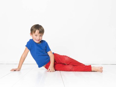 altı yaşındaki yakışıklı kırmızı pantolon ve gülümseyen ve beyaz katta otururken kameraya bakarak mavi t-shirt