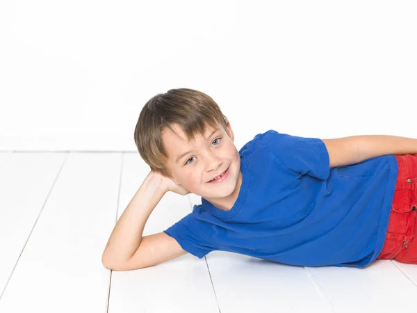 6岁男孩 身穿红色长裤和蓝色 躺在白色木地板上微笑着看着相机 — 图库照片