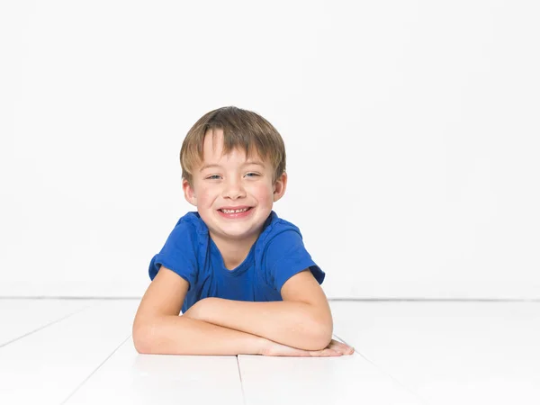 6岁的男孩在蓝色 T恤看着相机 躺在白色的地板交叉的武器 — 图库照片