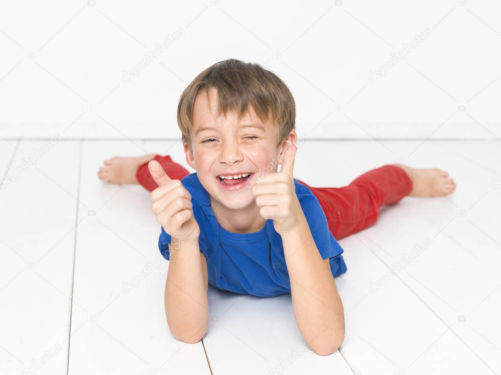 six year old boy in red trousers and blue t-shirt laughing and showing thumbs up while lying on white wooden floor