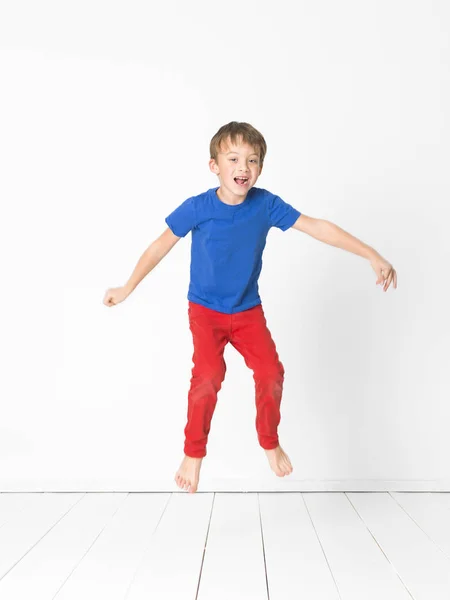 スタジオで白い壁の前に木製の床を飛び越えて高青の シャツと赤のズボンのかわいい男の子 — ストック写真
