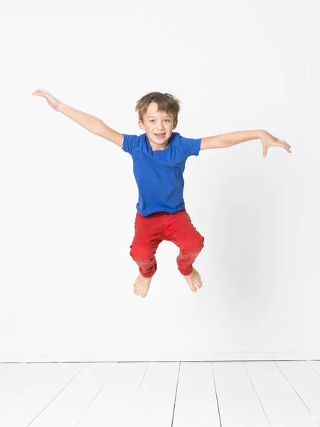 スタジオで白い壁の前に木製の床を飛び越えて高青の シャツと赤のズボンのかわいい男の子 — ストック写真